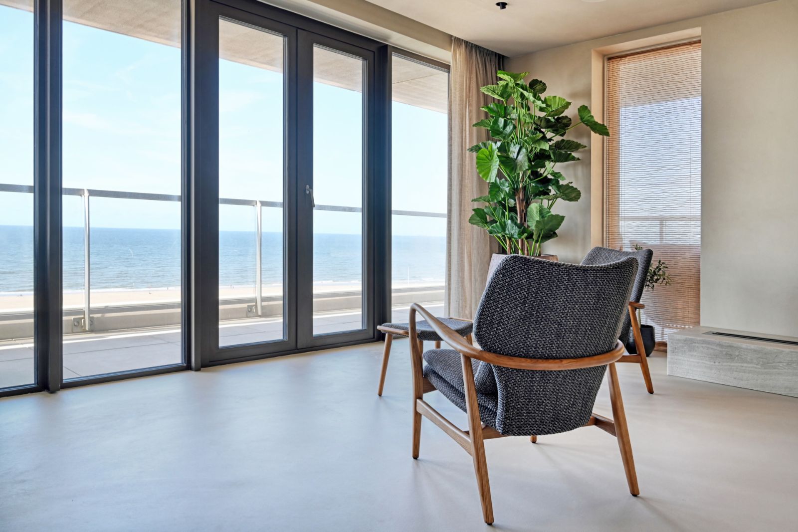 Woonkamer met uitzicht op zee en een lichtgrijze Pava gietvloer, met een donkere fauteuil en een grote plant